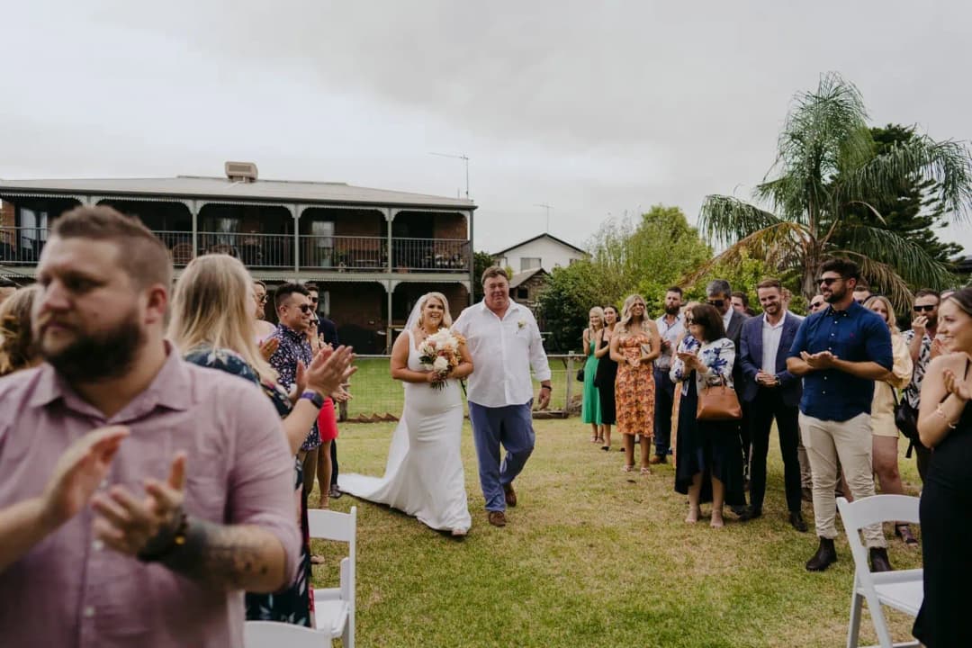 Bride entering the lakeside ceremony beside a man as guests watch