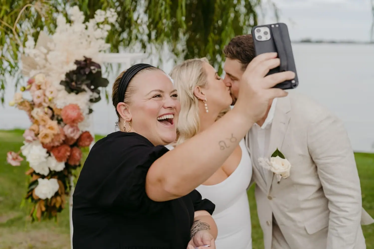 Celebrant taking a selfie while the bride and groom kiss behind her