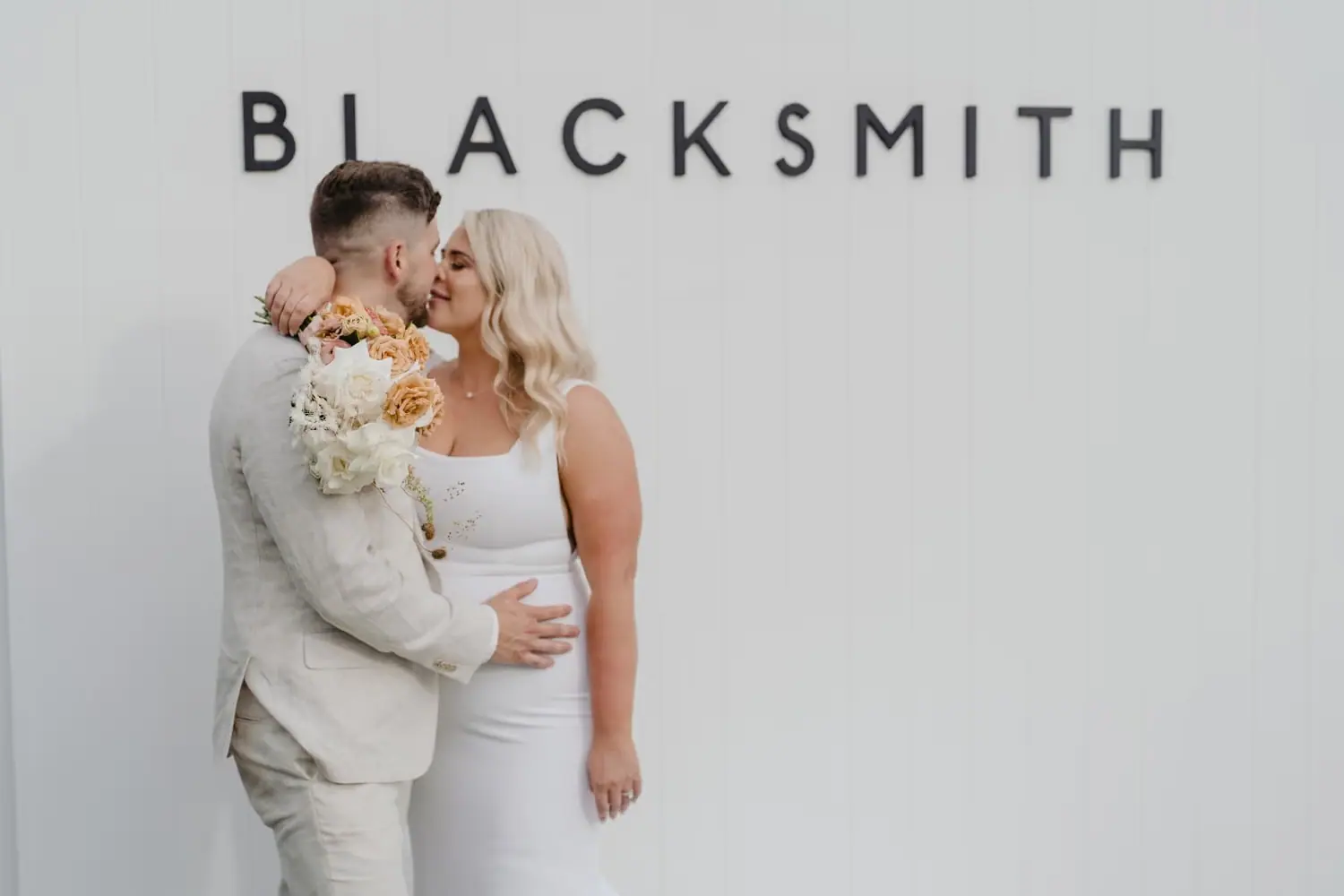 Bride and groom kissing beneath Blacksmith lettering on a white wall