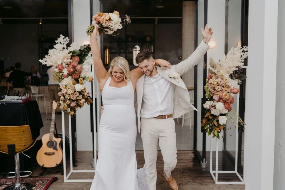 Bride raising her bouquet as the groom raises one arm beside the reception entrance