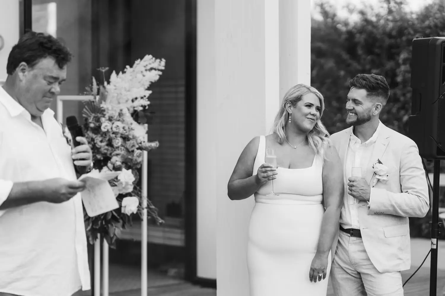 Black and white photograph of a guest reading from a paper beside the bride and groom