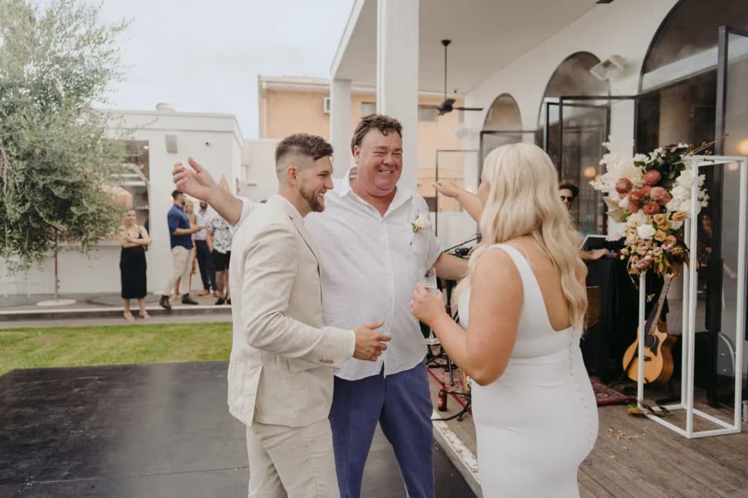 Bride and groom smiling as a man greets them with his arms open