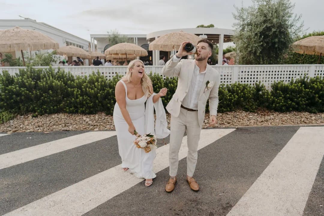 Bride laughing as the groom drinks from a bottle on a paved path