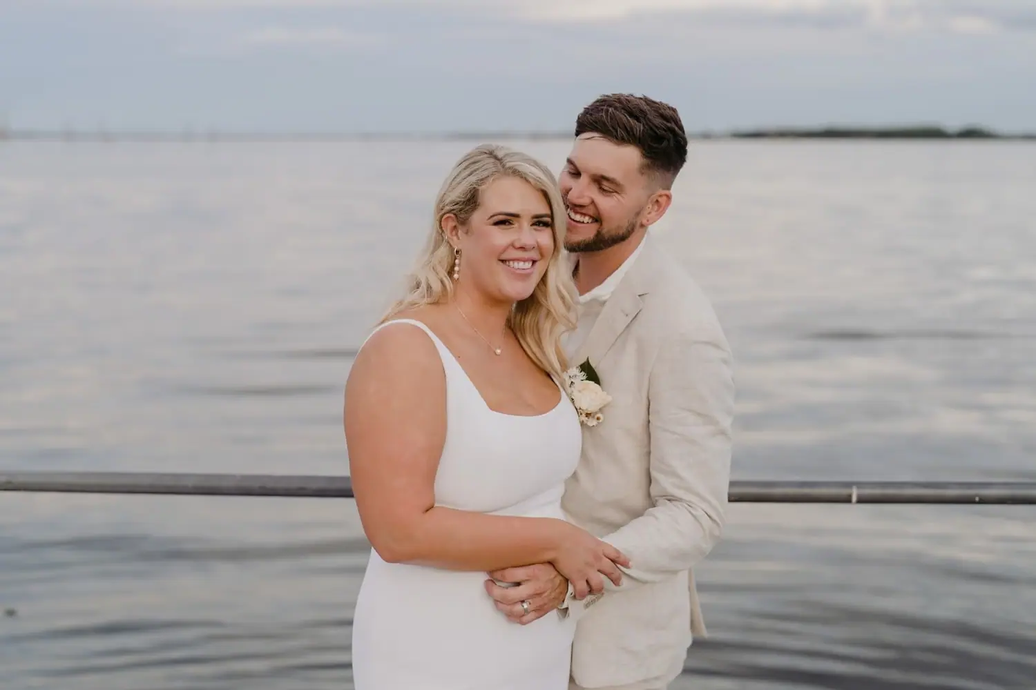 Bride smiling as the groom embraces her from behind beside the water
