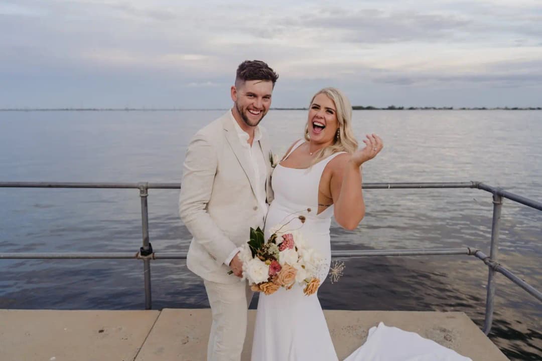 Bride and groom smiling toward the camera with a bouquet on a jetty