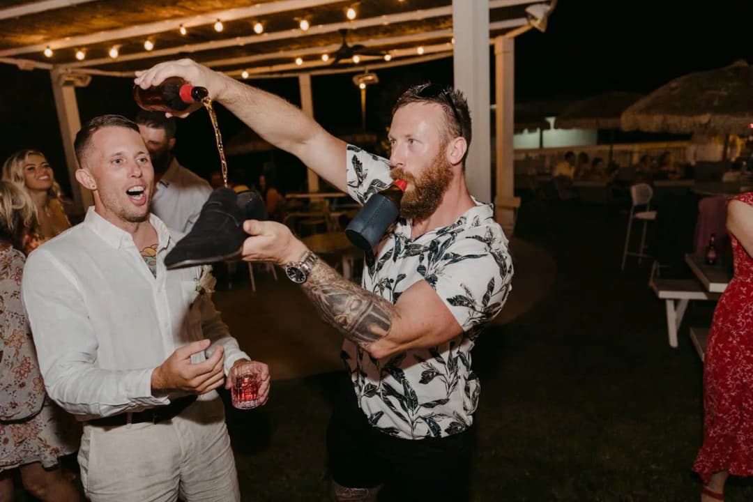 Guest pouring a drink from a bottle into a shoe beside another laughing guest