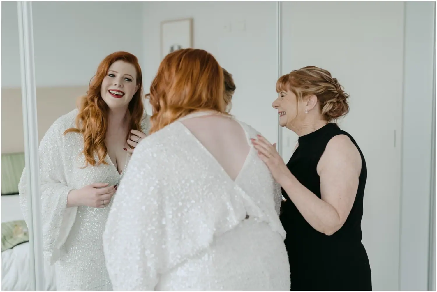 Bride smiling while a woman adjusts her sparkling wedding dress