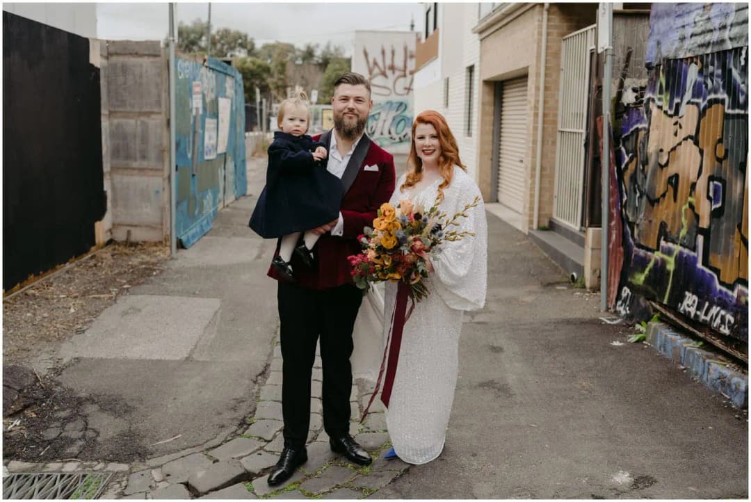 Groom holding a young child while standing beside the bride in a graffiti-lined laneway