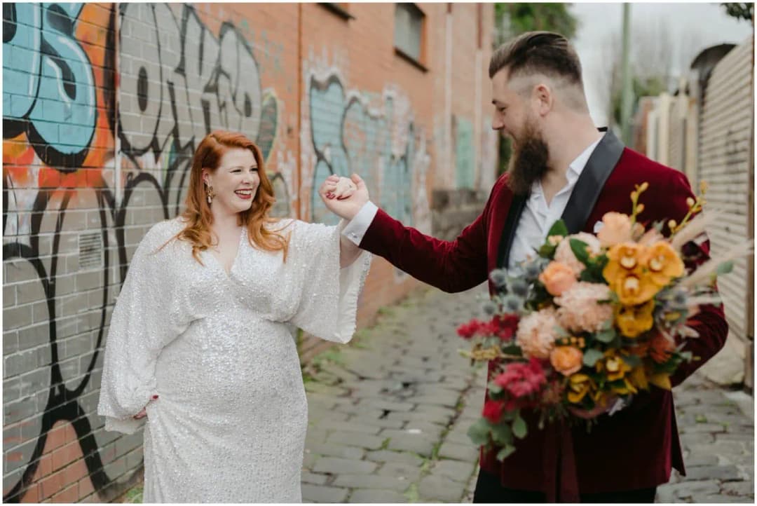 Groom raising the bride's hand as she smiles beside a graffiti-covered wall