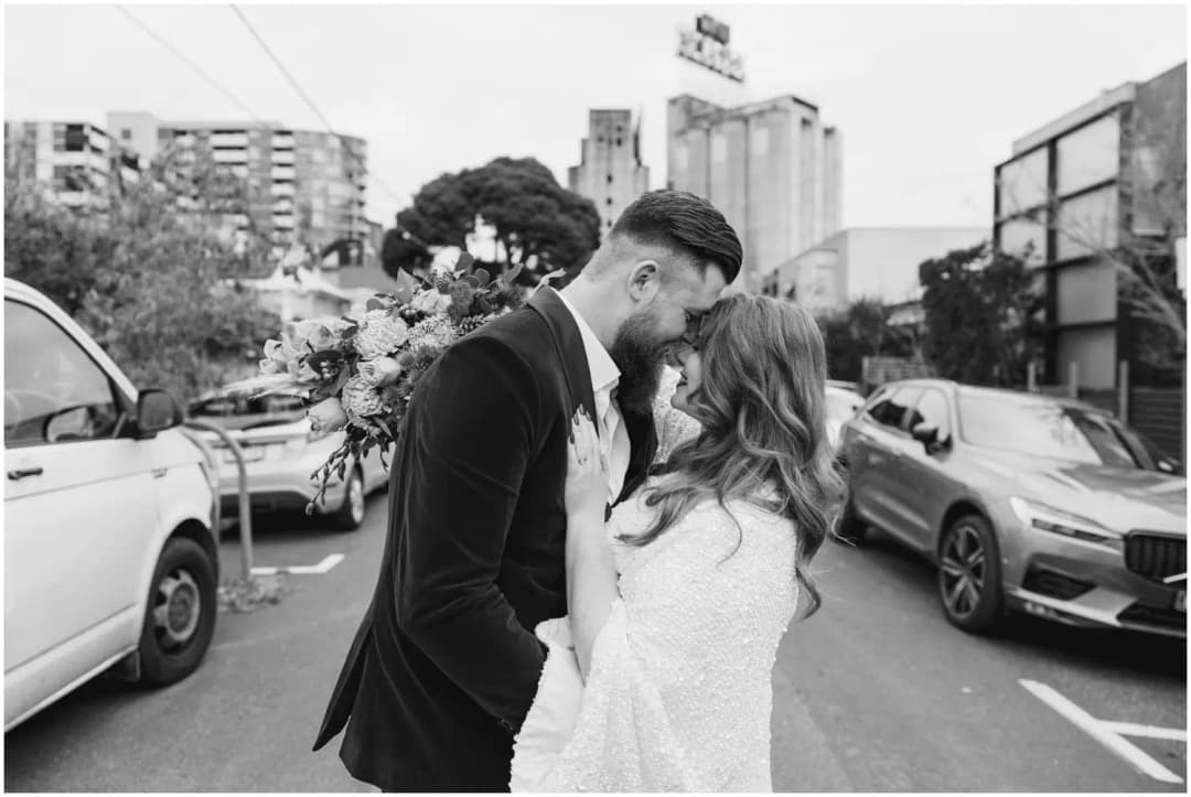 Black-and-white photograph of the bride and groom kissing in a parked-car-lined street