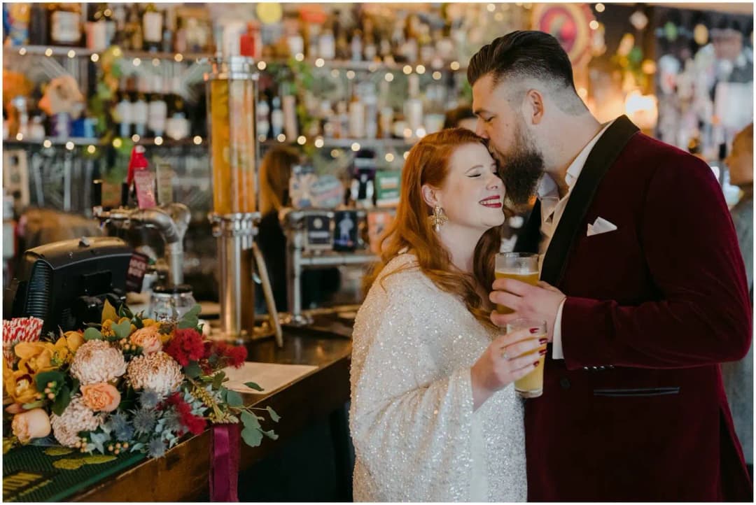 Groom kissing the bride's forehead while they hold drinks beside a pub bar