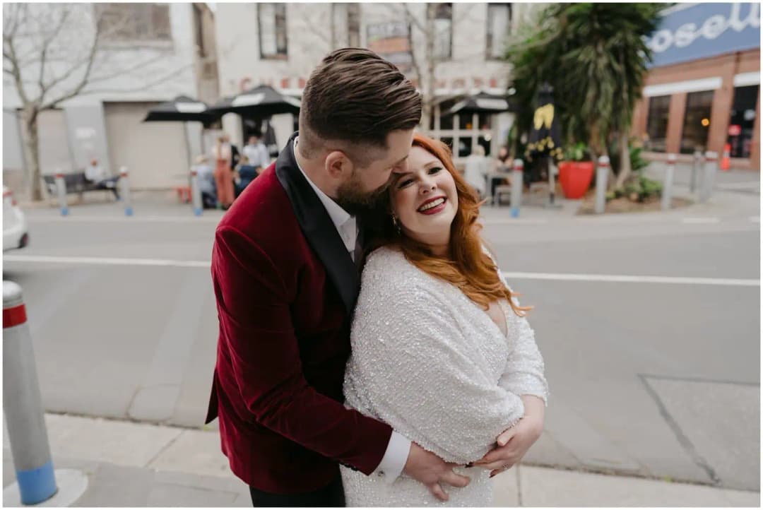 Groom embracing the laughing bride from behind beside a city street