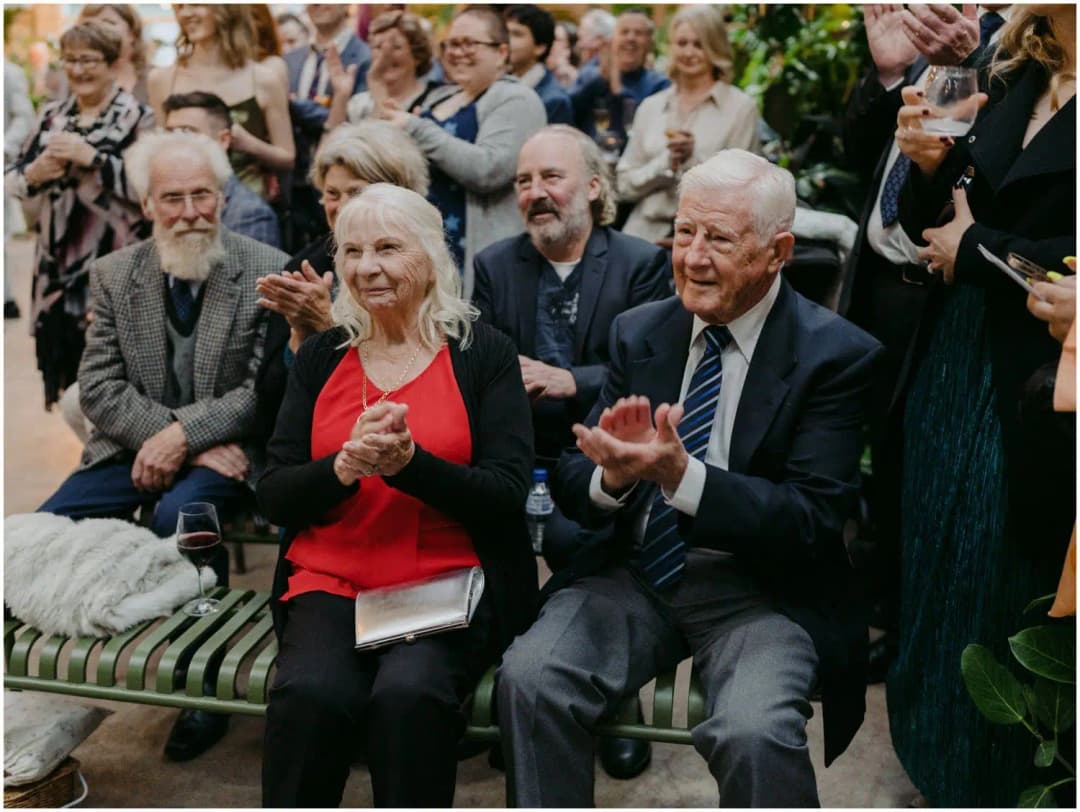 Seated wedding guests smiling and applauding inside a plant-filled venue