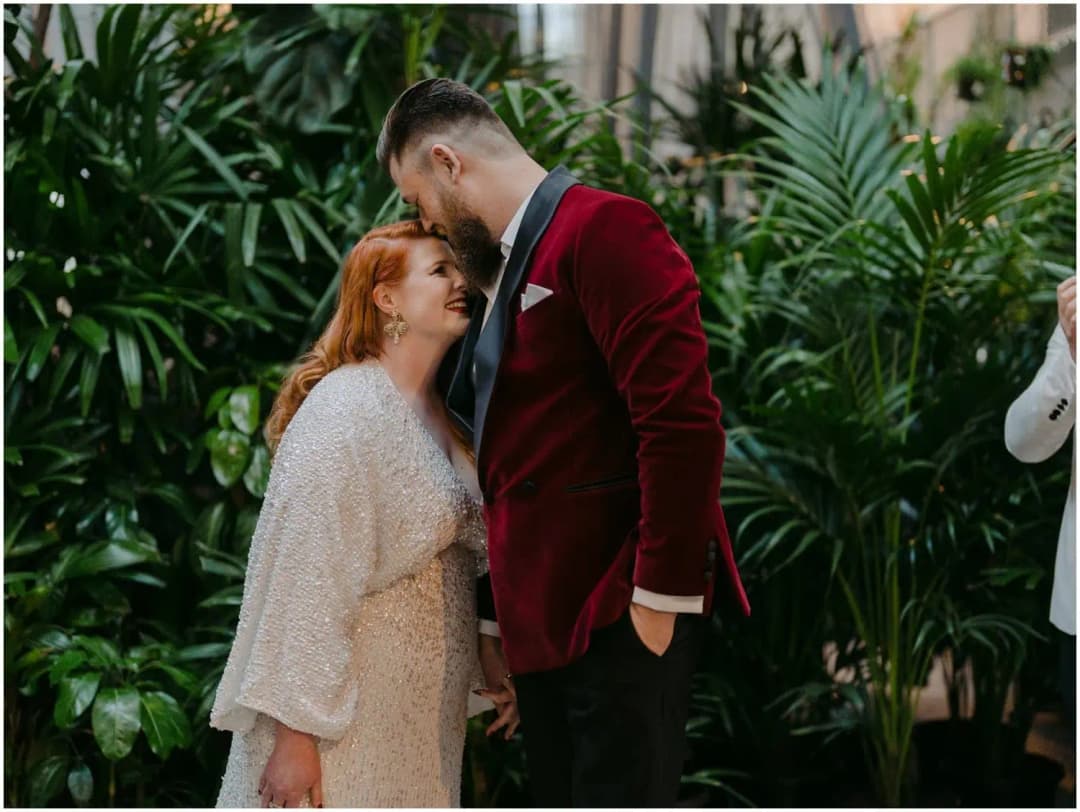 Groom leaning down to kiss the bride's forehead amid lush green plants