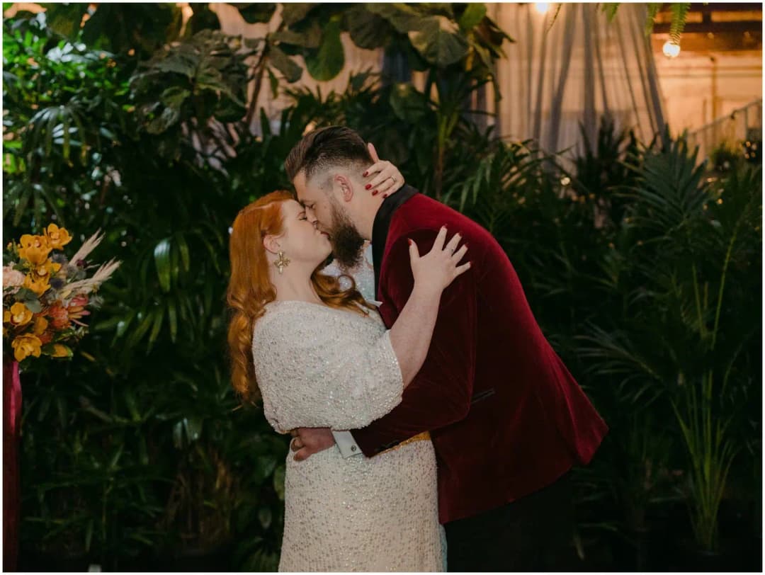 Bride and groom kissing in front of a wall of green plants