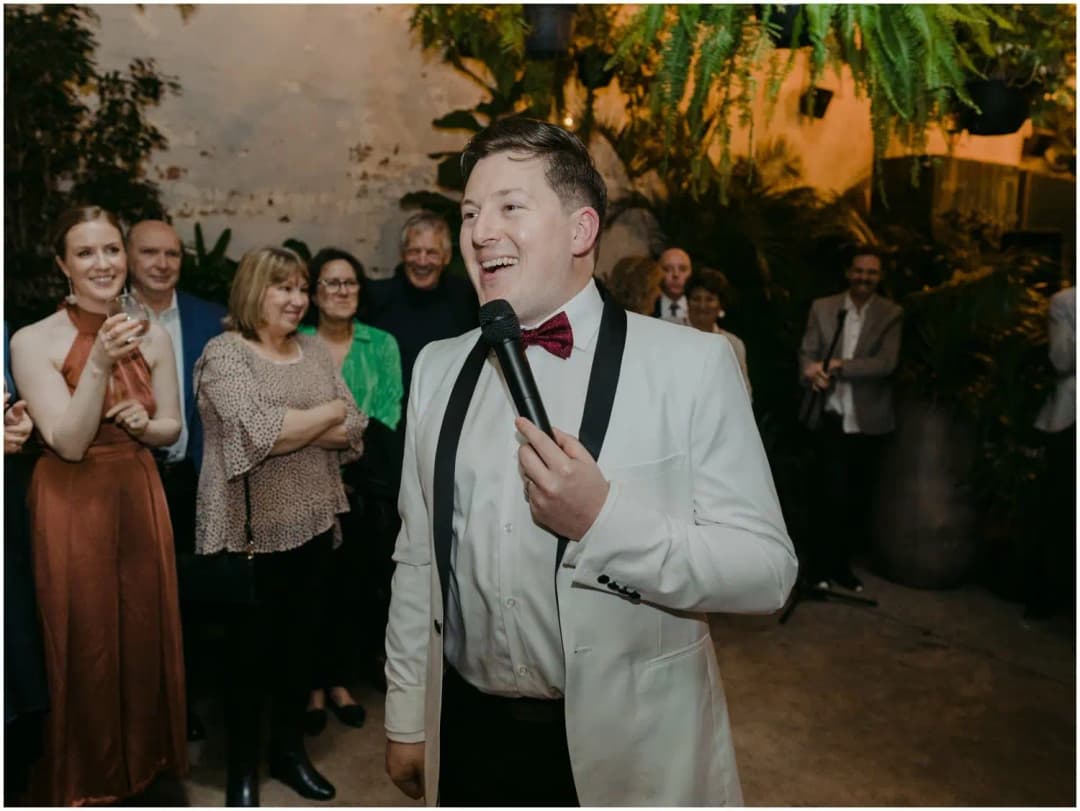 Man in a white jacket and bow tie speaking into a microphone while guests smile