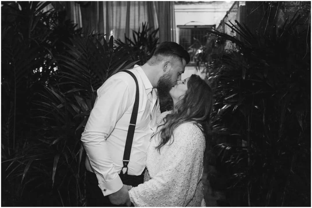 Black-and-white photograph of the bride and groom kissing amid leafy plants