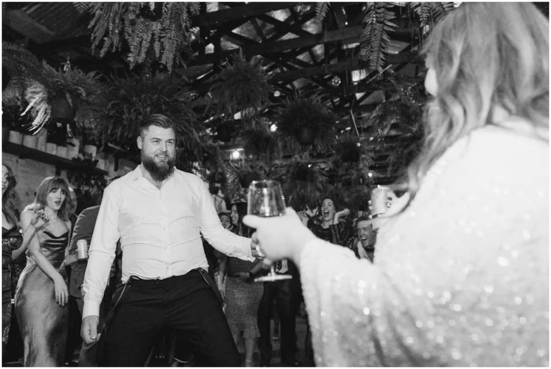 Black-and-white photograph of the groom dancing toward the bride as she holds a wine glass