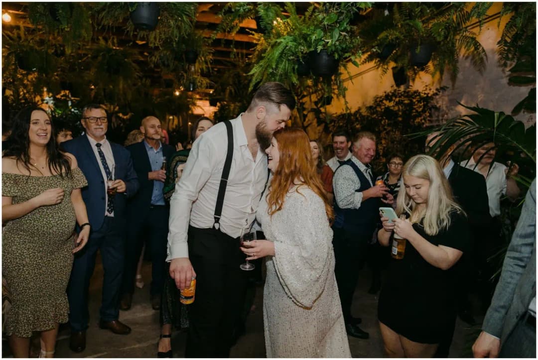 Bride and groom kissing among dancing guests beneath hanging plants