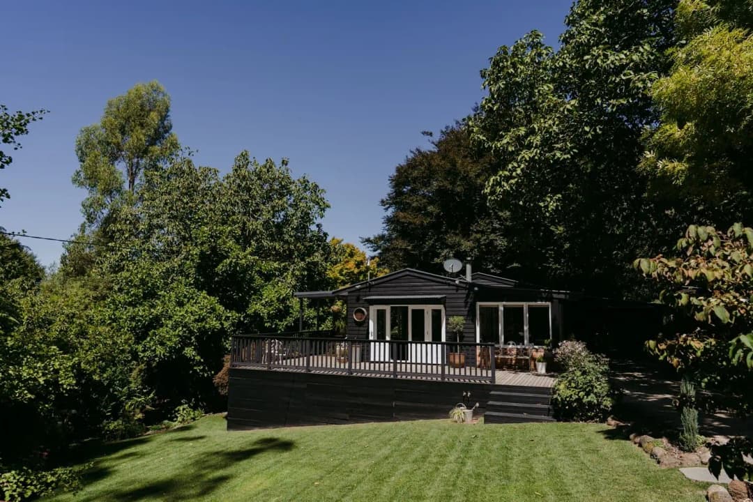 Dark timber cabin with a veranda overlooking a green lawn and leafy garden