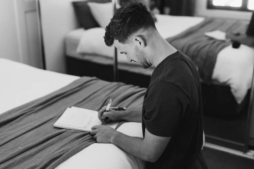 Black and white photograph of a man writing in a notebook beside a bed