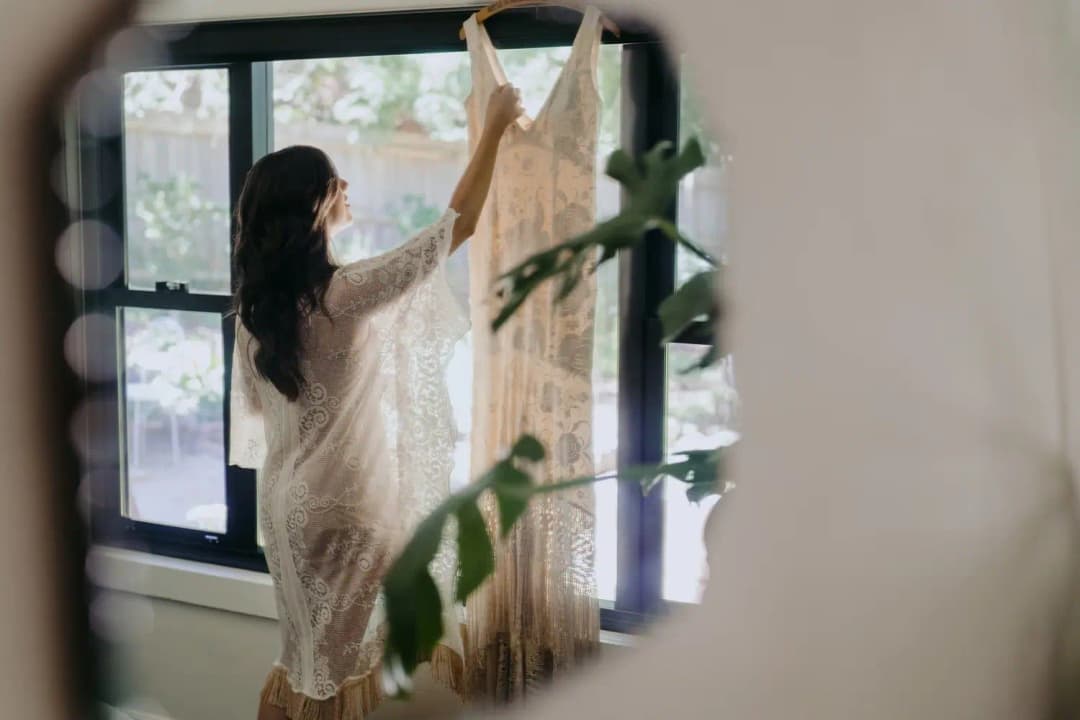 Bride holding her wedding dress up beside a window