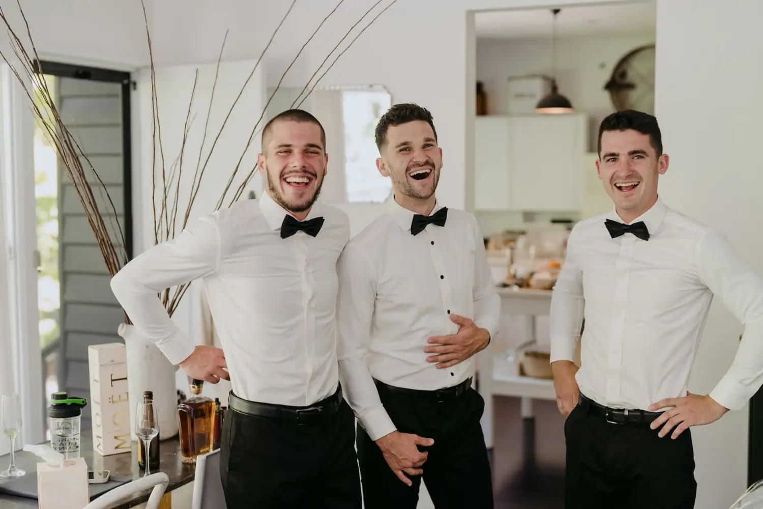 Three men in white shirts and black bow ties laughing together indoors