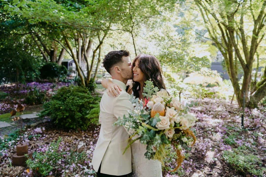 Bride and groom kissing while holding a pale flower bouquet in the garden