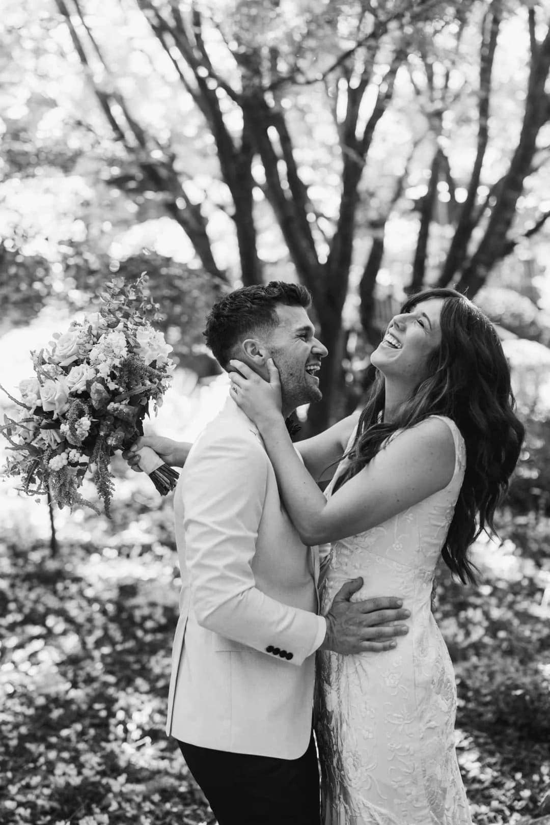 Black and white photograph of the bride and groom laughing together beneath trees