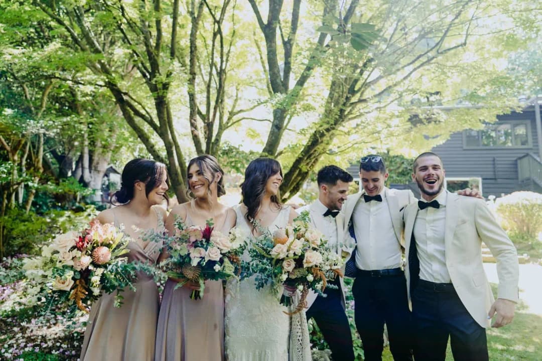 Wedding party laughing together with bouquets beneath leafy trees