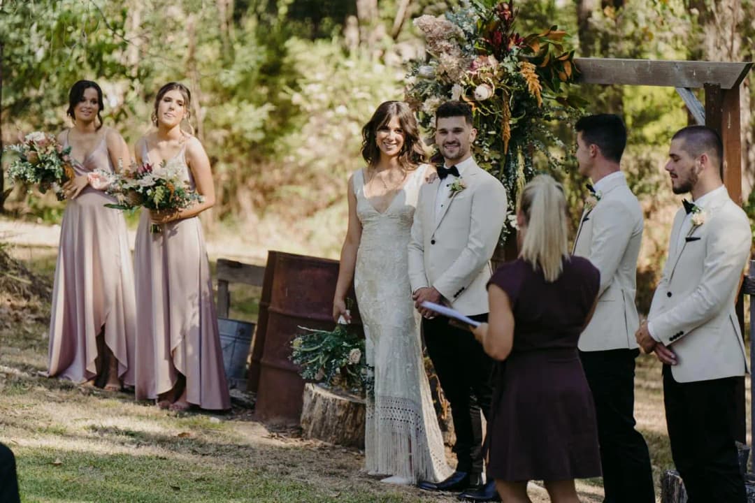 Bride and groom standing with their wedding party beside a rustic timber ceremony entrance