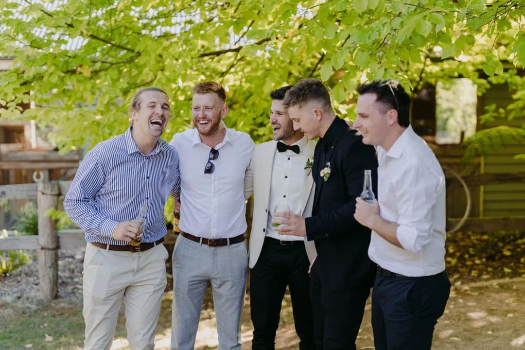 Groom and three guests laughing together with drinks beneath trees