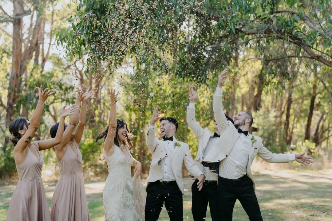 Wedding party cheering with raised arms as sparkling wine sprays beneath trees