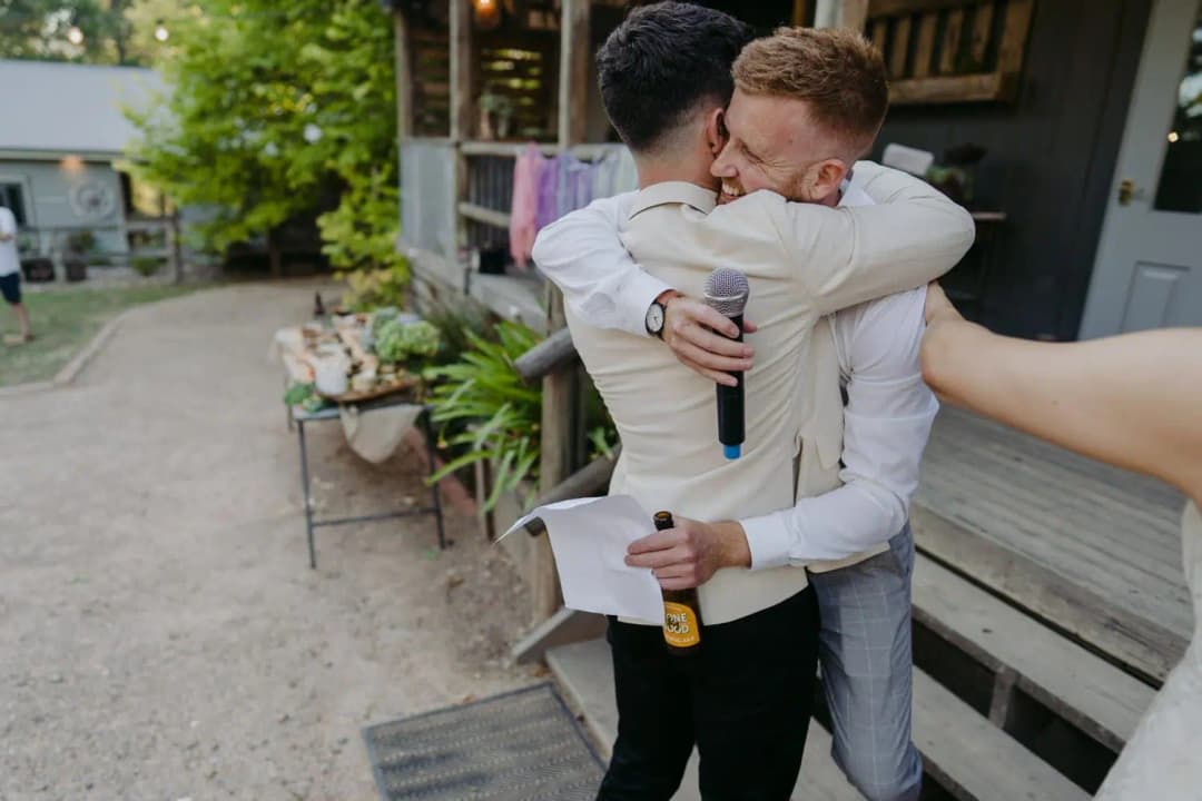 Groom hugging a guest who holds a microphone and speech notes