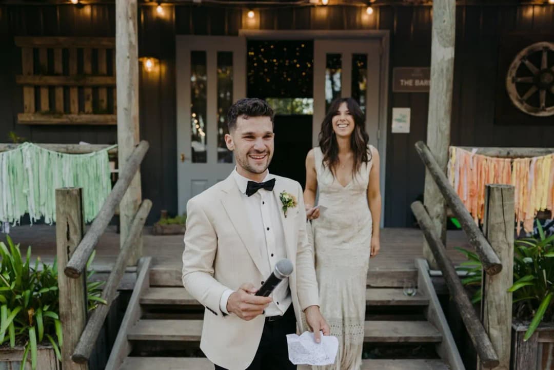 Groom speaking into a microphone with the smiling bride behind him on the steps