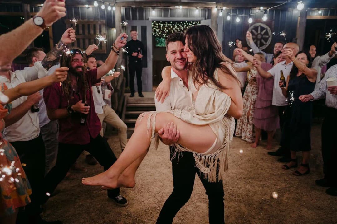 Groom carrying the smiling bride as guests raise drinks around them