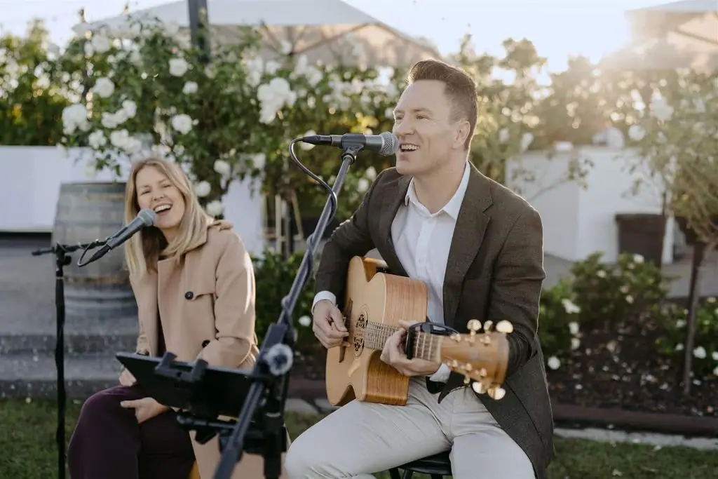 Singer and guitarist performing together outside beside wedding flowers.