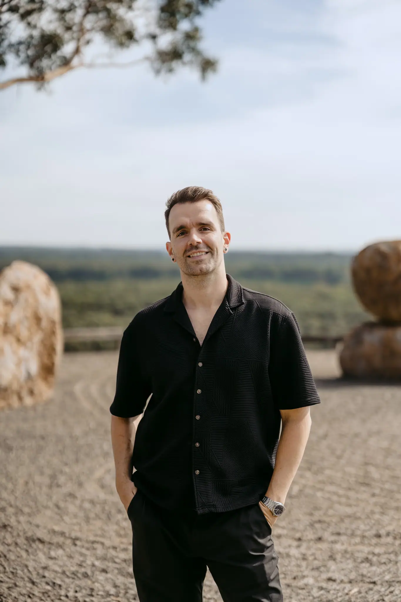 Man in a black shirt smiling beside stacked hay bales.