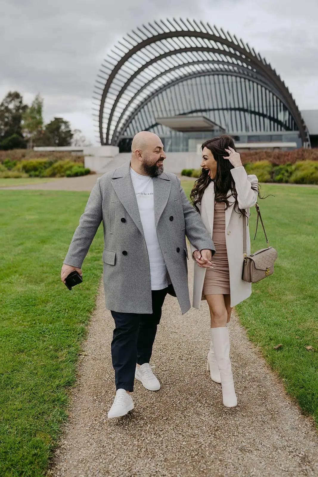 Couple walking hand in hand near a large curved-roof building.