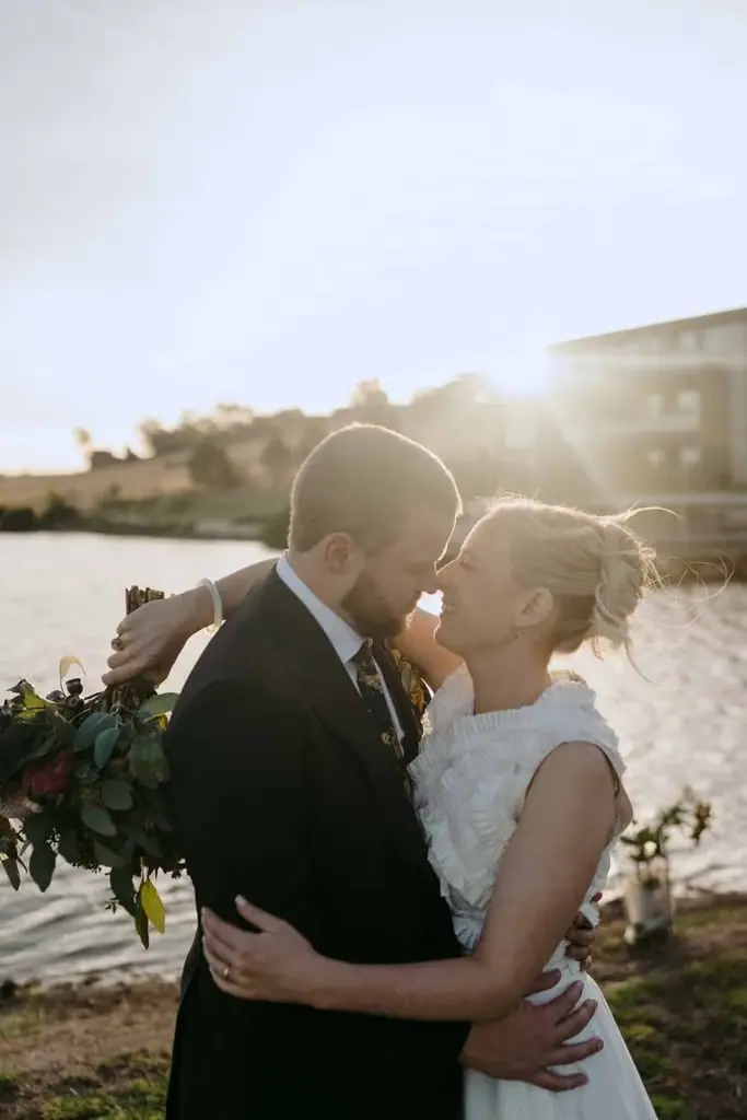 Groom kissing the bride's head beside the lake at sunset