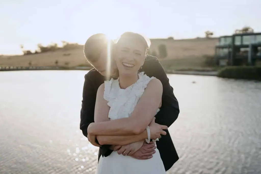 Groom embracing the smiling bride from behind beside the lake