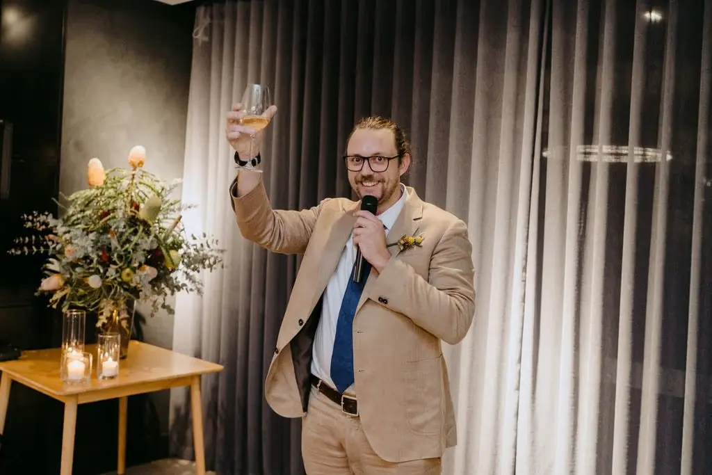 Guest in a tan suit raising an arm while speaking into a microphone