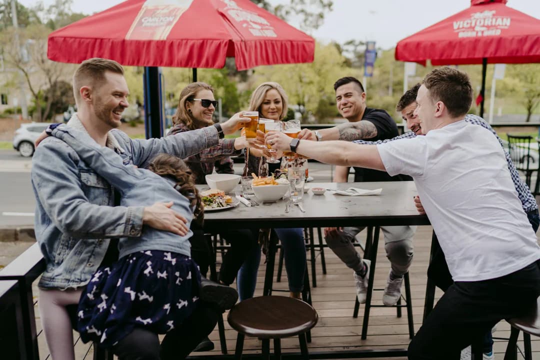 Guests clinking beer glasses around an outdoor table beneath red umbrellas.
