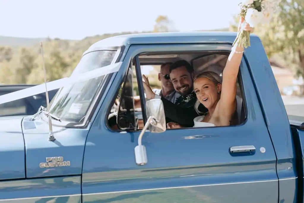 Newlyweds smiling from the cab of a blue pickup truck