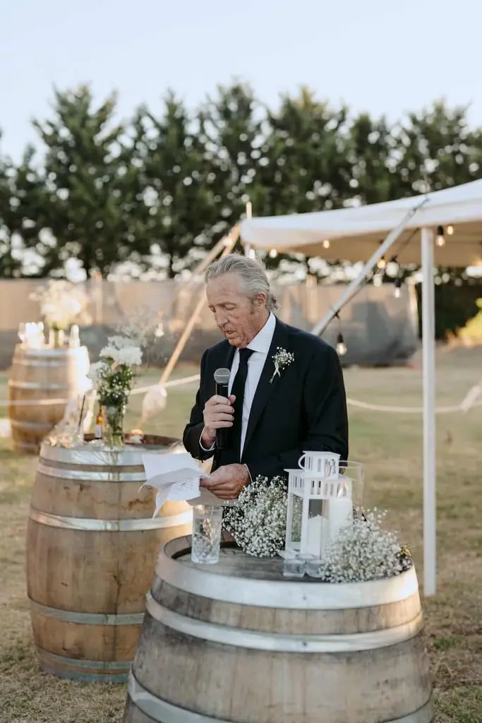 Older guest speaking into a microphone beside a wine-barrel table