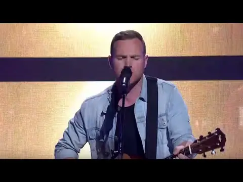 Guitarist singing into a microphone against a softly lit stage backdrop.