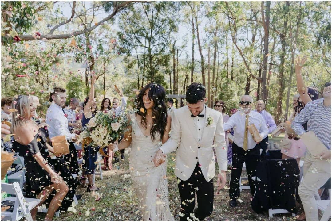 Couple walking through guests throwing pale confetti in a woodland clearing.