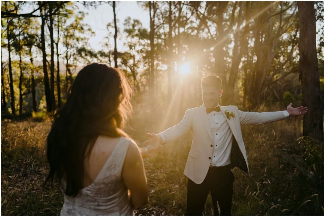 Groom spreading his arms toward the bride in golden forest light.