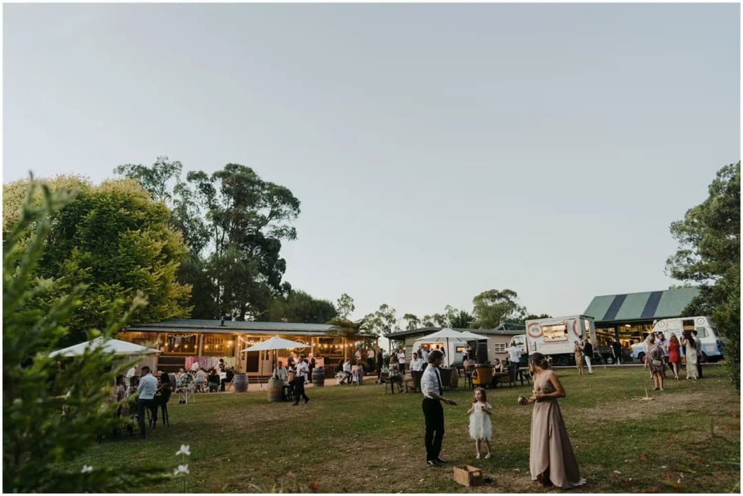 Guests socialising across a lawn beside timber buildings at dusk.