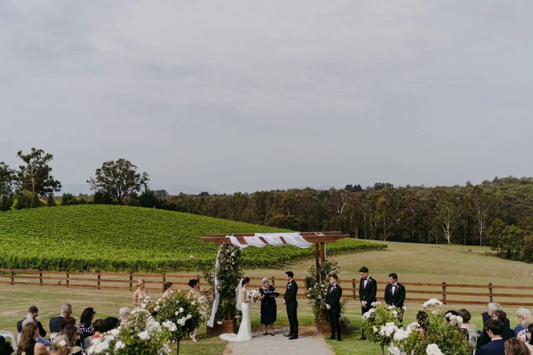 Outdoor ceremony beneath a floral arch overlooking grapevines and green fields.